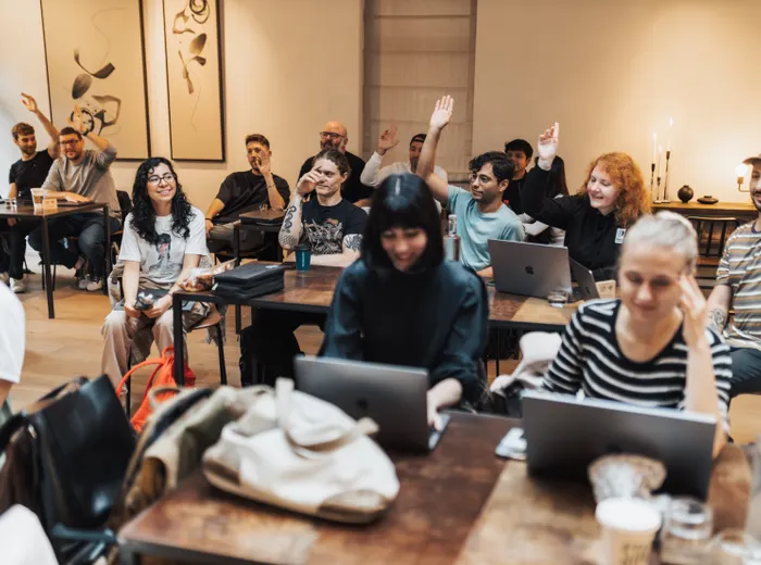 Uma sala cheia de gente sentada às mesas com notebooks; várias pessoas levantam a mão.