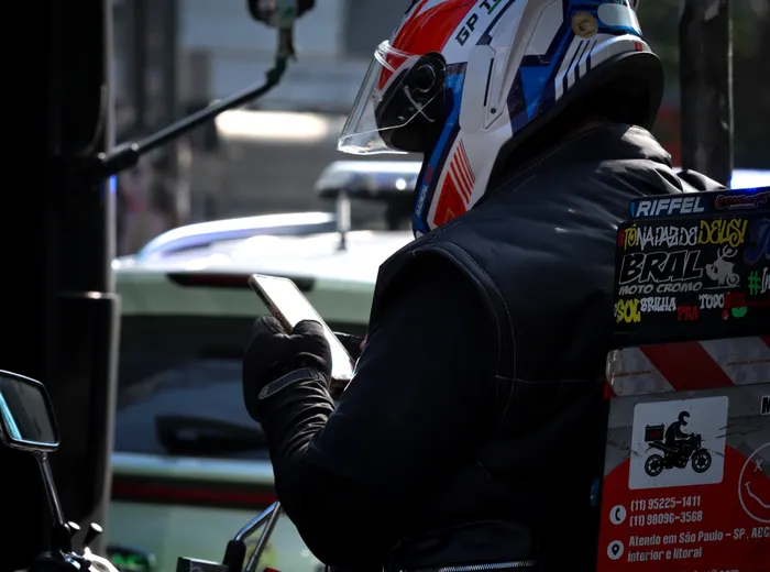 Motociclista de capacete e colete, parado no trânsito de São Paulo, olhando o celular.
