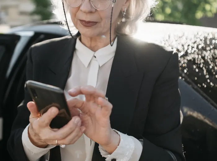 Mãos de uma mulher de terno mexendo no celular, encostada na porta de um carro, na rua.